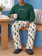 Man wearing green Merry Christmas long sleeve tee and Christmas tree pajama pants seated with cocoa