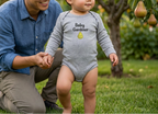 A baby wearing a light gray long-sleeve bodysuit with 'Baby Gardener' and a pear graphic stands on grass while holding an adult's hand in a pear orchard.
