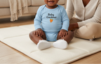 A baby wearing a light blue long-sleeve bodysuit with 'Baby Gardener' and a small orange carrot graphic sits on a cream floor mat in a bright living room, next to a smiling woman.