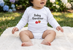 A baby sitting outdoors on a cream knit blanket, wearing a white long-sleeve bodysuit with the text 'Baby Gardener' and a small strawberry graphic. The background shows green grass and blurred hydrangea flowers.