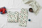 Folded Blue Christmas men’s pajama top and pocketless pants on white bedding with cocoa, a book, greenery, and a gift