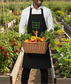 A man wearing a black apron with green text that reads 'Grow it Cook it Love it' holds a wicker basket filled with fresh vegetables in a sunny garden.