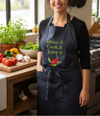 A woman wearing a navy blue kitchen apron with the text 'Grow it Cook it Love it' and a red and green chili pepper graphic, standing in a bright, vegetable-filled kitchen.