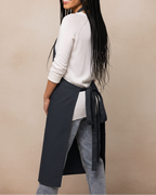 A woman wearing a dark gray kitchen apron over a white long-sleeve top and gray jeans, shown from a side-back angle against a plain beige studio background.