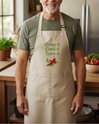 A smiling older man wearing a beige apron over an olive green t-shirt in a warm kitchen setting. The apron features the printed text 'Grow it Cook it Love it' in green, with a graphic of a red chili pepper and green leaves.