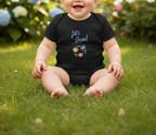 A baby sitting outdoors on green grass wearing a black short-sleeve bodysuit with the text 'Let's Grow!' and small colorful flower graphics on the chest.