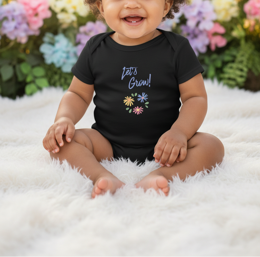 A smiling baby sitting cross-legged on a white fluffy rug, wearing a black short-sleeve bodysuit with the text 'Let's Grow!' and a small colorful flower graphic on the chest. The background shows blurred flowers and greenery.