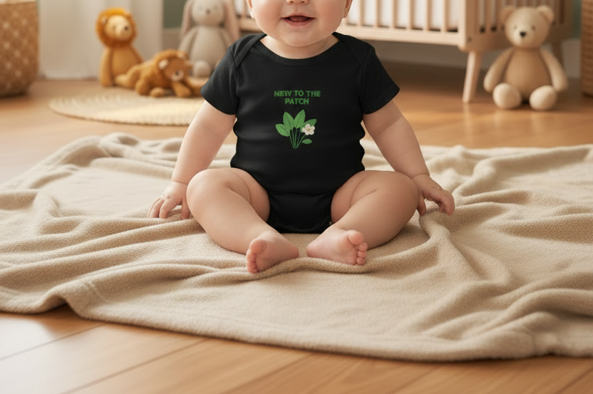 A baby sitting on a beige blanket on a wooden floor in a nursery, wearing a black short-sleeve bodysuit with green text reading 'NEW TO THE PATCH' and a green leaf graphic.