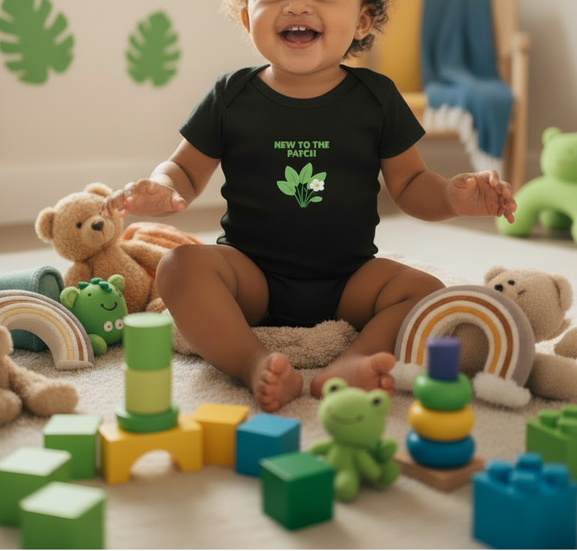 A baby sitting on a carpeted floor surrounded by colorful toys, wearing a black short-sleeve bodysuit with a green graphic that reads 'NEW TO THE PATCH!' and a small leaf illustration.