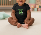 A baby sitting cross-legged on a light beige carpet wearing a black short-sleeve bodysuit with green text reading 'NEW TO THE PATCH' and a green leaf graphic with a small white flower.