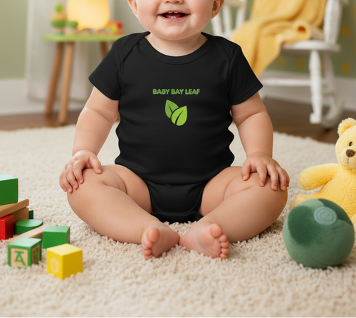 A baby sitting on a cream carpet in a playroom, wearing a black short-sleeve bodysuit with a green leaf graphic and the text 'BABY BAY LEAF'.