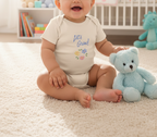 A baby sitting on a cream carpet next to a light blue teddy bear, wearing a cream bodysuit with the text 'Let's Grow!' and small colorful flowers. A crib and toy shelf are visible in the softly blurred background.