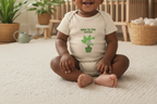 A smiling baby sitting cross-legged on a cream textured rug in a nursery, wearing a beige short-sleeve bodysuit with a green leaf graphic and the text 'NEW TO THE PATCH'.