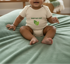 A baby sitting on a soft green blanket wearing a cream bodysuit with 'BABY BAY LEAF' and a green leaf logo printed on the chest.