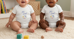 Two babies sitting on a cream carpet wearing matching white bodysuits with a blue 'Let's Grow!' graphic and small floral print.