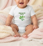 A smiling baby wearing a white short-sleeve bodysuit with a green 'NEW TO THE PATCH' graphic and leaf illustration, sitting on a soft cream and pink blanket.