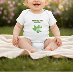 A baby sitting outdoors on a blanket wearing a white short-sleeve bodysuit with green text reading 'NEW TO THE PATCH' and a green leaf-and-flower graphic.