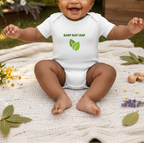 A baby sitting on a white knit blanket outdoors, wearing a white bodysuit with green text reading 'BABY BAY LEAF' and a green leaf graphic on the chest.