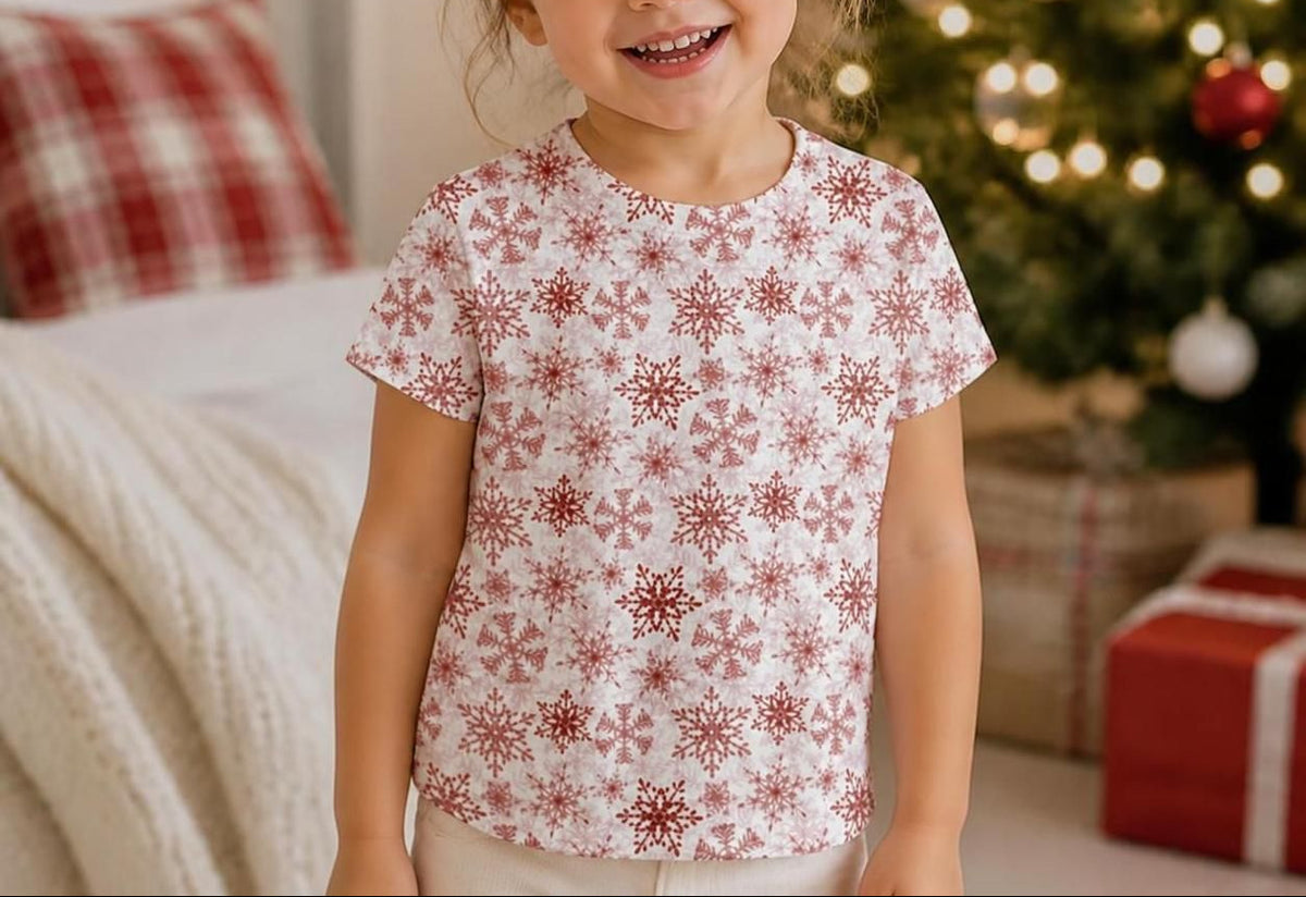 A young girl smiling in a holiday-themed bedroom, wearing a white short-sleeve t-shirt with a red snowflake pattern. A decorated Christmas tree, wrapped gifts, and a cozy bed are visible in the background.