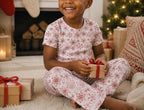 A young child sitting on a rug in a cozy holiday living room, wearing a matching short-sleeve pajama set with a red snowflake pattern on a white background, holding a small gift box with a red ribbon.