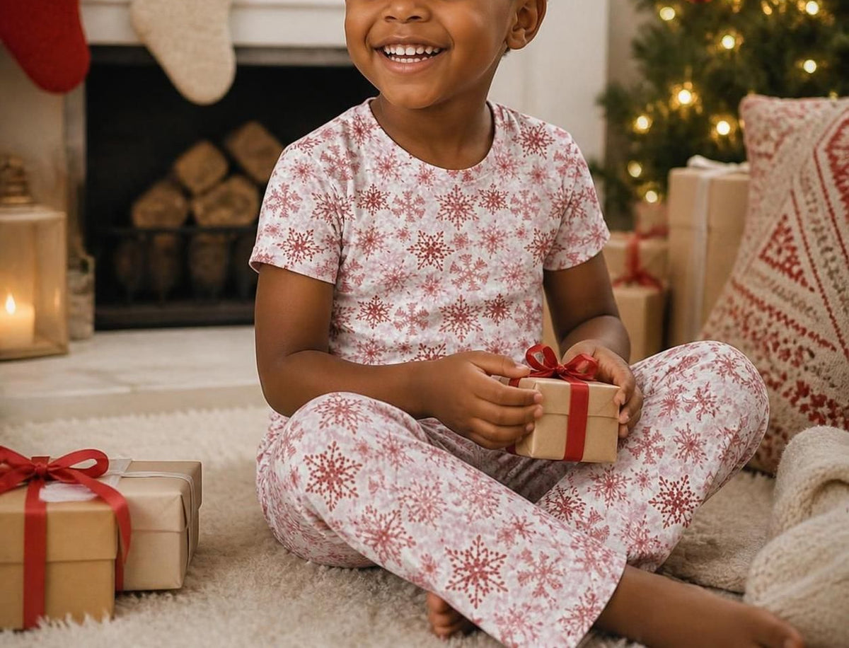 A young child sitting on a rug in a cozy holiday living room, wearing a matching short-sleeve pajama set with a red snowflake pattern on a white background, holding a small gift box with a red ribbon.