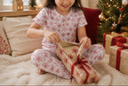 A young girl with dark hair sits cross-legged on a fluffy cream rug, opening a gift box wrapped in beige paper with a red snowflake pattern and a red ribbon. She is wearing matching pajamas with a red snowflake pattern.