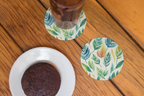 Coaster with leaf pattern next to a chocolate cookie and a glass of iced coffee on a wooden surface.