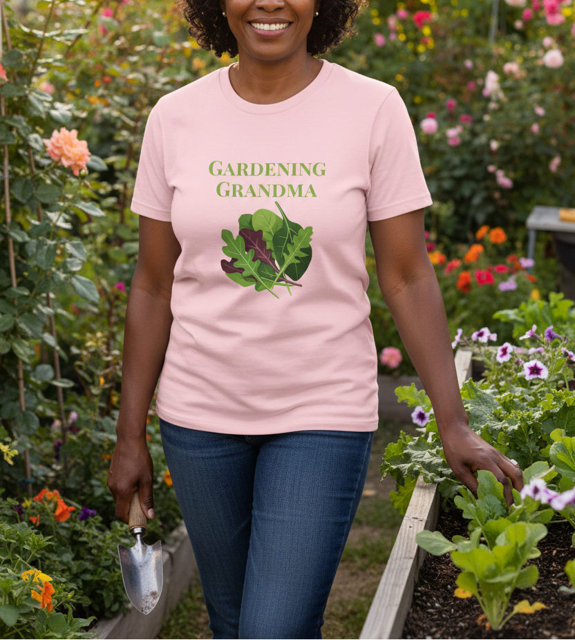 A woman wearing a light pink t-shirt with a green graphic that reads 'GARDENING GRANDMA' above an illustration of leafy greens, standing in a lush garden.