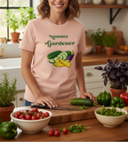 A woman wearing a light pink graphic t-shirt with the text 'Mommy Gardener' and a vegetable illustration, standing in a kitchen behind a wooden cutting board with fresh vegetables.