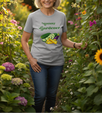A woman wearing a light gray graphic t-shirt that says 'Mommy Gardener' with vegetable illustrations, standing in a lush garden.