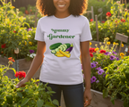 A woman wearing a white graphic t-shirt that reads 'Mommy Gardener' with vegetables, standing in a sunny garden.