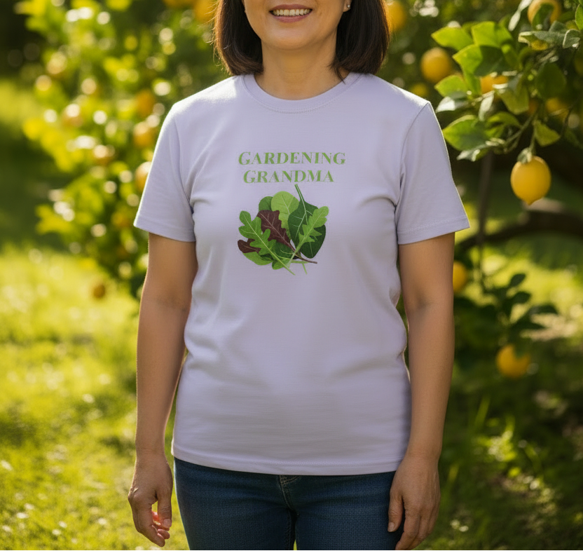 A woman wearing a light lavender graphic t-shirt with the words 'GARDENING GRANDMA' and a green leaf illustration, standing in a sunny garden with lemon trees in the background.